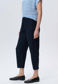 Personne portant un pantalon noir coupé avec des ourlets retroussés, un haut tricoté bleu clair à manches courtes, et des chaussures plates noires, se tenant devant un fond uni.