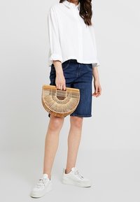 Bolso de bambú tejido con un diseño semicircular, sostenido por una persona que lleva una camisa blanca y pantalones cortos de mezclilla, combinados con zapatillas blancas.