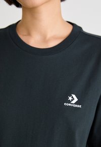 Melna kokvilnas T-kreklā ar apaļu kakla un īsām piedurknēm. Uz krūtīm ir balts "Converse" logo ar zvaigznes un bultas dizainu.