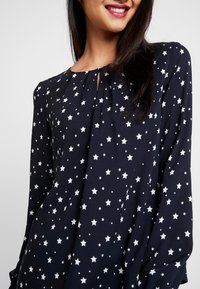 Blusa blu navy con maniche lunghe, caratterizzata da un motivo a stelle bianche. Dettaglio con scollatura a chiave e tessuto morbido e fluido.