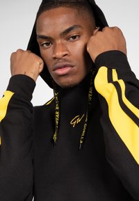 Zwarte hoodie met gele strepen, voorzien van een trekkoord met logo tekst. Materiaal lijkt textuur te hebben; nauwsluitend ontwerp.