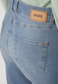 Jean en denim bleu clair avec une texture lisse, comprenant une pièce en cuir étiquetée "ANGELS" et un petit bouton en métal près de la poche arrière.