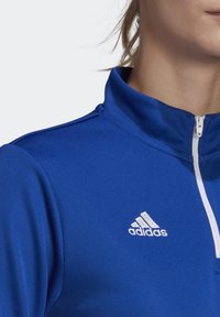 Chaqueta atlética azul con cremallera y cuello alto, que presenta una cremallera blanca y un logotipo bordado en blanco de Adidas en el pecho izquierdo.