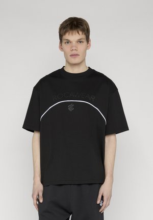 Fiatal férfi rövid barna hajjal, fekete Rocawear túlméretezett pólóban, melyen fehér, ívelt vonal fut a mellkasán keresztül.