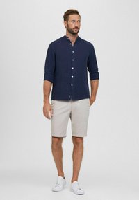 Camicia di lino blu navy con colletto a banda, maniche arrotolate; pantaloni corti beige; sneakers bianche. Design semplice, vestibilità su misura, tessuto morbido.