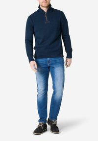 Marineblau gestrickter Pullover mit einem Viertelreißverschluss und hohem Kragen, kombiniert mit hellblauen Jeans und schwarzen Wildlederschuhen. Auffälliges Etikett am Ärmel.
