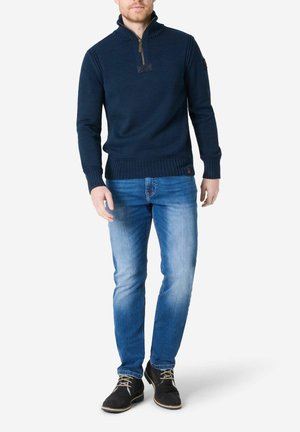 Marineblau gestrickter Pullover mit einem Viertelreißverschluss und hohem Kragen, kombiniert mit hellblauen Jeans und schwarzen Wildlederschuhen. Auffälliges Etikett am Ärmel.