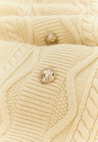 Maglione lavorato a maglia color crema con motivi testurizzati, trecce e dettagli a costine, con bottoni metallici per la chiusura.
