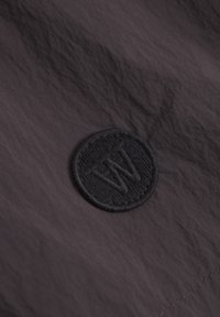 Tissu noir texturé avec un logo circulaire brodé présentant des lettres stylisées. Le matériau semble léger et durable.