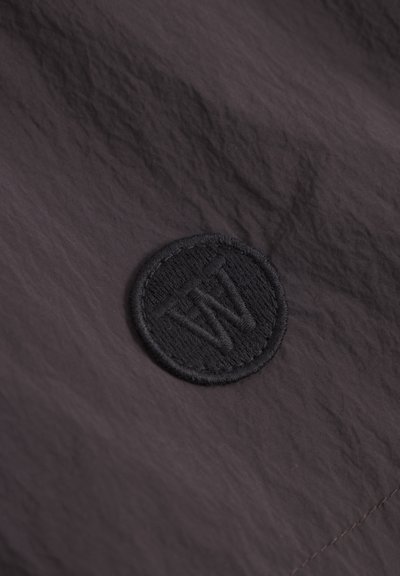 Tissu noir texturé avec un logo circulaire brodé présentant des lettres stylisées. Le matériau semble léger et durable.