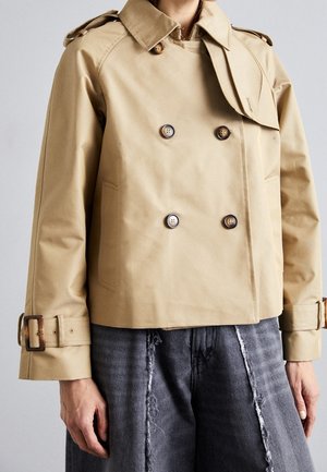 Trench-coat beige court croisé avec épaulettes, poignets bouclés, porté avec un jean gris délavé à jambes larges et usées.
