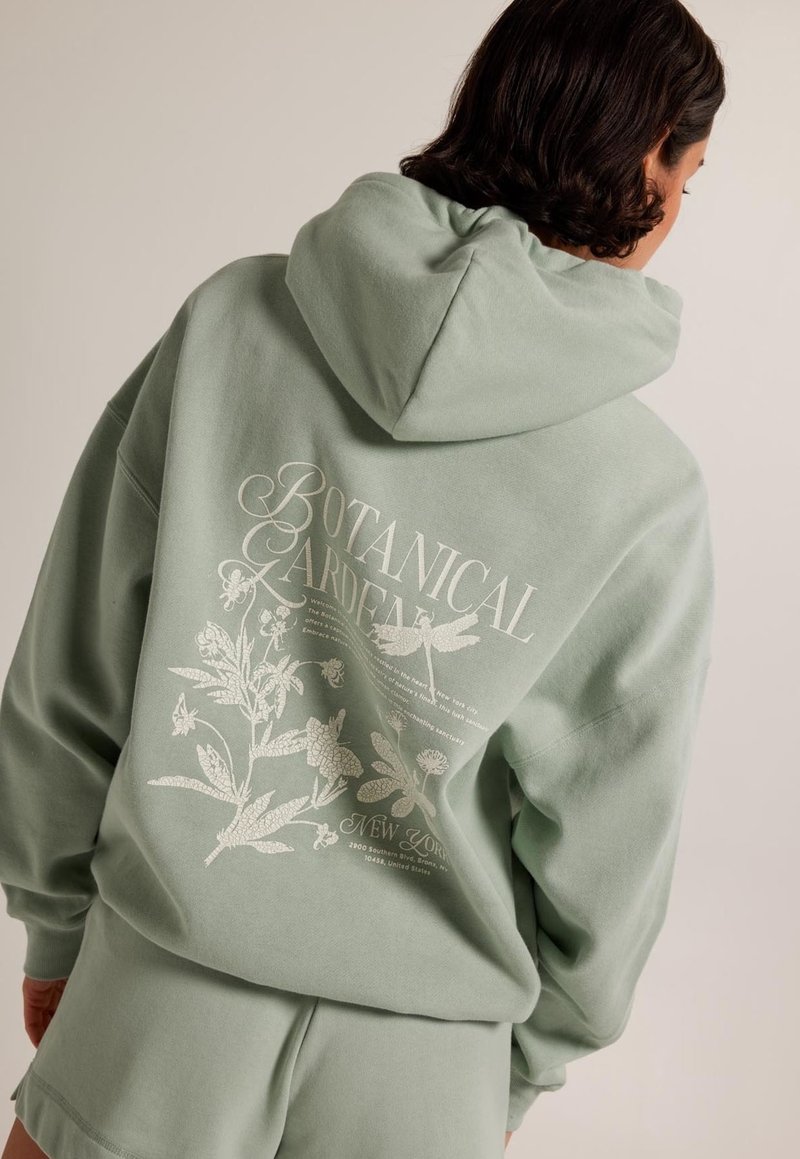 Personne portant un sweat à capuche vert sauge et un short, le dos du sweat à capuche montrant une impression botanique florale blanche et le texte "Jardin Botanique New York".