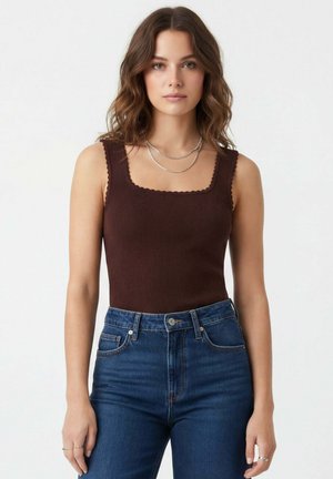 Jeune femme aux cheveux bruns ondulés portant un haut côtelé sans manches de couleur marron foncé et un jean taille haute bleu, sur un fond blanc uni.