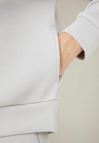 Felpa grigia chiaro con tasca laterale, polsini a coste e texture liscia e morbida. Il tessuto ha una leggera lucentezza.