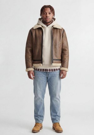 Homme portant une veste en peau lainée marron par-dessus un sweat à capuche beige et une chemise à carreaux, jean bleu clair et bottes couleur sable, debout devant un fond uni.
