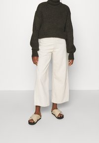 Pullover a collo alto chunky di colore verde scuro abbinato a pantaloni larghi di colore crema e sandali color nocciola con suola nera e cinghie incrociate.