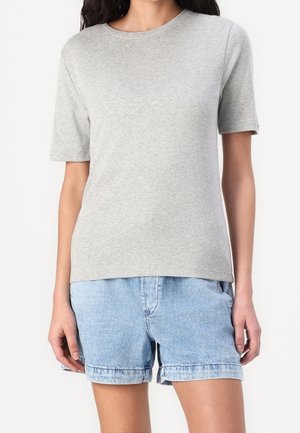 Kvinde iført en ensfarvet lysegrå T-shirt med korte ærmer og lyseblå denimshorts mod en hvid baggrund.