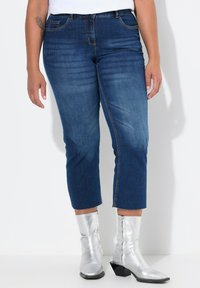 Tummansiniset lyhyet farkut, joissa on kevyt kulutus, viisi taskua ja riipustettu helma, yhdistettynä metallinhohtoisiin hopeisiin nilkkabootseihin.