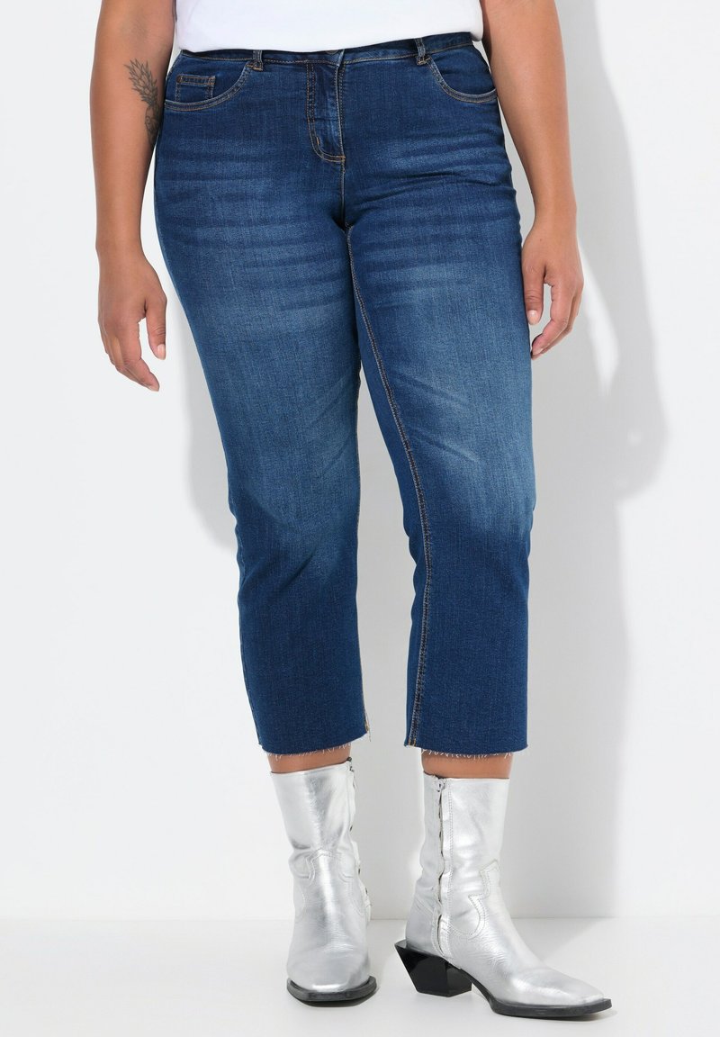 Tummansiniset lyhyet farkut, joissa on kevyt kulutus, viisi taskua ja riipustettu helma, yhdistettynä metallinhohtoisiin hopeisiin nilkkabootseihin.