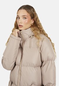 Kurtka puchowa w jasnym beżowym kolorze, z wysokim kołnierzem, zapięciem na zamek z przodu, sznurkiem przy szyi oraz pikowaną fakturą z bocznymi kieszeniami.