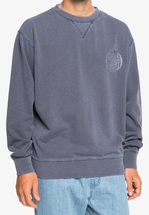 Mann trägt einen ausgewaschenen dunkelgrauen Sweatshirt mit Rundhalsausschnitt, kleinem, kreisförmigem, geprägtem Logo auf der Brust und hellblauen Jeans.