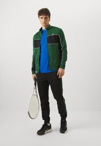 Groene en zwarte zip-up sportjack met witte piping, blauwe t-shirt, zwarte joggingbroek met groene zijstreep en zwarte sportschoenen. Houdt een tennisracket vast.