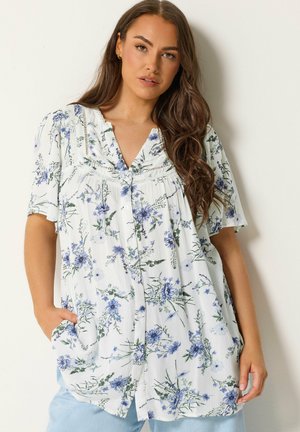 Kvinde med langt brunt hår iført en hvid kortærmet bluse med blå blomsterprint og lyseblå bukser, står mod en hvid væg.