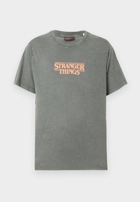 Dark green cotton T-shirt with short sleeves, featuring a peach "STRANGER THINGS" logo in retro font centered on the chest.