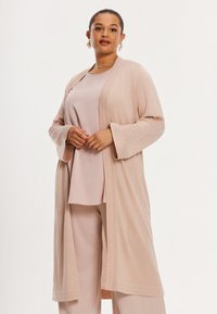 Langer, offen frontierter Cardigan aus hellrosa strukturiertem Stoff, mit weiten Ärmeln und lässiger Passform, getragen über einem passenden Top und einer Hose.