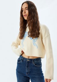 Maglione cropped color crema con design grafico blu, maniche lunghe e texture morbida, abbinato a jeans scuri a vita alta.