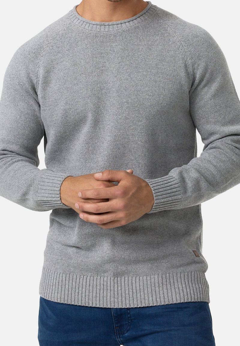 Homme portant un pull en maille gris clair ajusté avec des poignets et un ourlet côtelés, mains jointes devant, associé à un jean bleu.