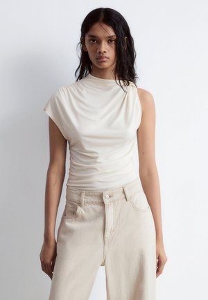Cream-colored sleeveless top with a draped design and gathered texture, paired with light beige high-waisted denim trousers.