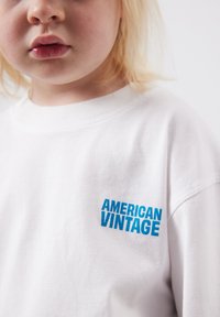 Jeune enfant aux cheveux blonds portant un t-shirt blanc avec le texte bleu "AMERICAN VINTAGE" sur la poitrine, sur un fond uni.