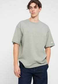 Tricou din bumbac verde deschis, cu mâneci scurte, cu croi lejer, având un branding subtil pe piept și un logo pe mânecă. Asortat cu blugi închisi la culoare.