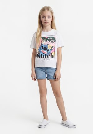 Jeune fille aux longs cheveux blonds portant un t-shirt blanc avec un motif Stitch, un short en jean et des chaussures blanches sans lacets, debout devant un fond blanc.