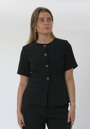 Jonge vrouw draagt een zwart kort mouw jasje met knopen en bijpassende broek, kijkt iets naar rechts tegen een effen achtergrond.
