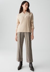 Beige gebreide trui met een V-hals, gecombineerd met grijs geruite hoge taille broek en spitse zwarte enkellaarsjes. Geweven stofdetails zichtbaar.