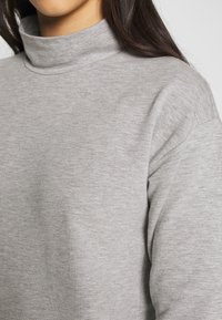 Světle šedý rolákový svetr s jemnou texturou, volným střihem a širokými manžetami. Má minimální švy a vysoký límec.