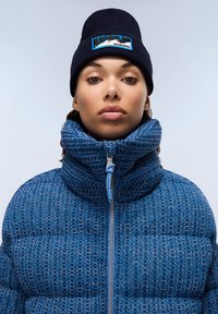 Blue puffer jacket with a high collar and textured pattern. Features a zipper closure and a light blue drawstring. Paired with a navy beanie.