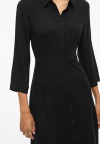 Juodas suknelė su mygtukų užsegimu priekyje, ilgomis rankovėmis ir prigludusia juosmeniu. Pagaminta iš lygaus audinio su matinio paviršiaus apdaila.