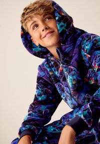 Paarse hoodie met kleurrijke abstracte patronen, getextureerd fleece materiaal, vooraan een rits en ribbelboorden. Heeft een grote capuchon en levendige accenten.