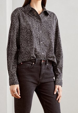 Femme portant une chemise noire à boutons avec des taches blanches, rentrée dans un jean noir taille haute, debout devant un fond clair uni.