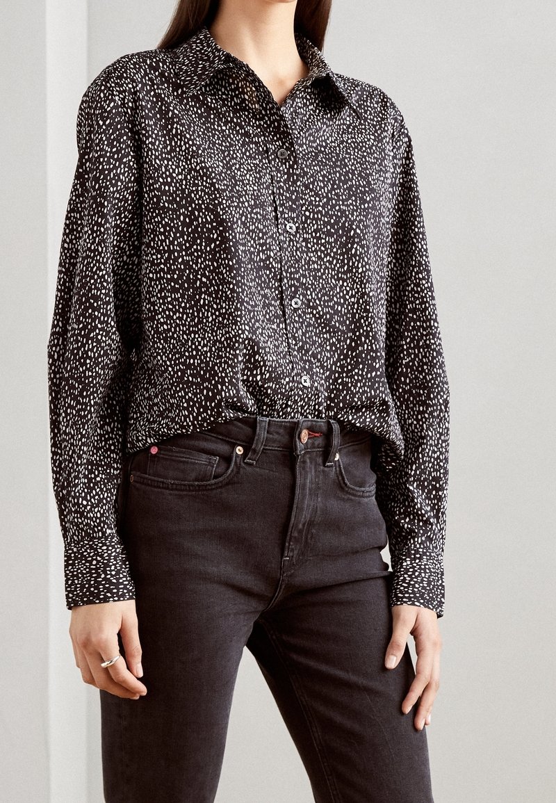 Femme portant une chemise noire à boutons avec des taches blanches, rentrée dans un jean noir taille haute, debout devant un fond clair uni.