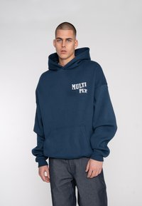 Dark blue hooded sweatshirt with front pocket and "MULTI PLY" printed logo in white. Soft texture and relaxed fit. Worn with dark jeans.