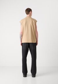 Beige bezrukávový top z měkkého materiálu, spárovaný s volnými černými kalhotami. Pohled zezadu ukazuje minimální švy a uvolněný střih.
