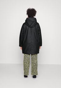 Schwarzer wasserdichter Mantel mit Kapuze, kombiniert mit floral gemusterten Hosen in Gelb und Schwarz, getragen mit schwarzen Schuhen.