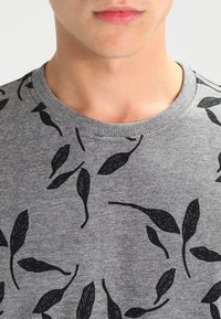 Sweat-shirt gris à col rond, avec un motif de feuilles noires. Le tissu semble doux avec une finition texturée autour du col.