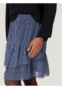 Jupe marine à couches avec un ourlet à volants, confectionnée en tissu léger avec un petit motif géométrique bleu et blanc. Taille élastique.