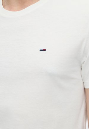 Ung mand iført en hvid T-shirt, mørkeblå jeans, hvide sneakers, en sølvkæde og et armbånd, stående med den ene hånd i lommen.