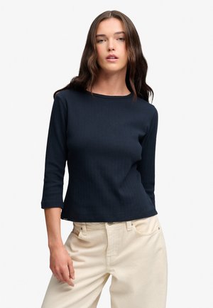 Vrouw met lang bruin haar, gekleed in een marineblauwe top met driekwart mouwen en crèmekleurige broek tegen een effen witte achtergrond.